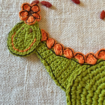 Close-up of the green crochet dinosaur head with an orange flower and embroidered face