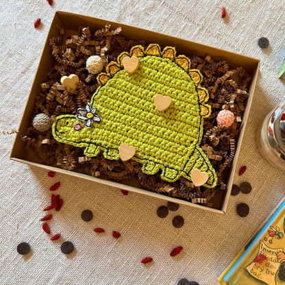 Green crochet dinosaur coaster with flower presented in a kraft gift box with decorative wooden hearts.