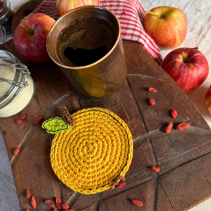 A round yellow crocheted coaster with a small green leaf accent and brown stem detail, styled on a dark wooden board with apples, a ceramic mug, and goji berries.