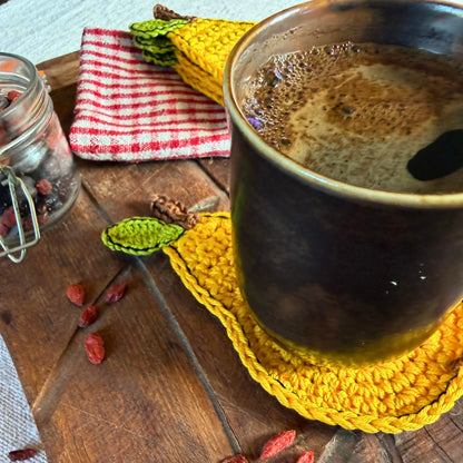 A dark ceramic mug filled with a hot coffee  sits on a yellow crocheted coaster on a rustic wooden table. A matching yellow crocheted leaf-shaped coaster and a red-and-white checkered cloth napkin are also visible, along with scattered goji berries and a glass jar.