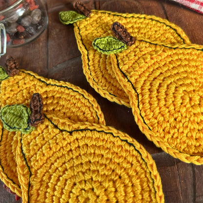 Four yellow crocheted pear-shaped coasters with green leaves and brown stems, arranged on a dark wooden surface.
