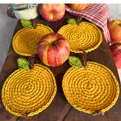 Four yellow crocheted apple - shaped coasters with green leaves and brown stems, arranged on a dark wooden board with whole apples and a red-and-white checkered cloth.