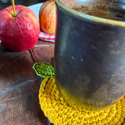 A close-up of a dark ceramic mug filled with coffee, resting on a yellow crocheted coaster on a wooden table. Two red apples are visible in the background.