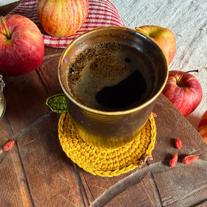 A ceramic mug filled with dark coffee sits on a yellow crocheted coaster on a rustic wooden table, surrounded by red apples and small red goji berries.