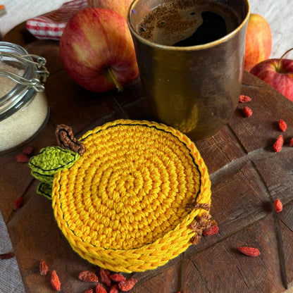 A round yellow crocheted coasters with a small green leaf and brown stem detail, styled on a rustic wooden board with apples, a cup of coffee, and goji berries.