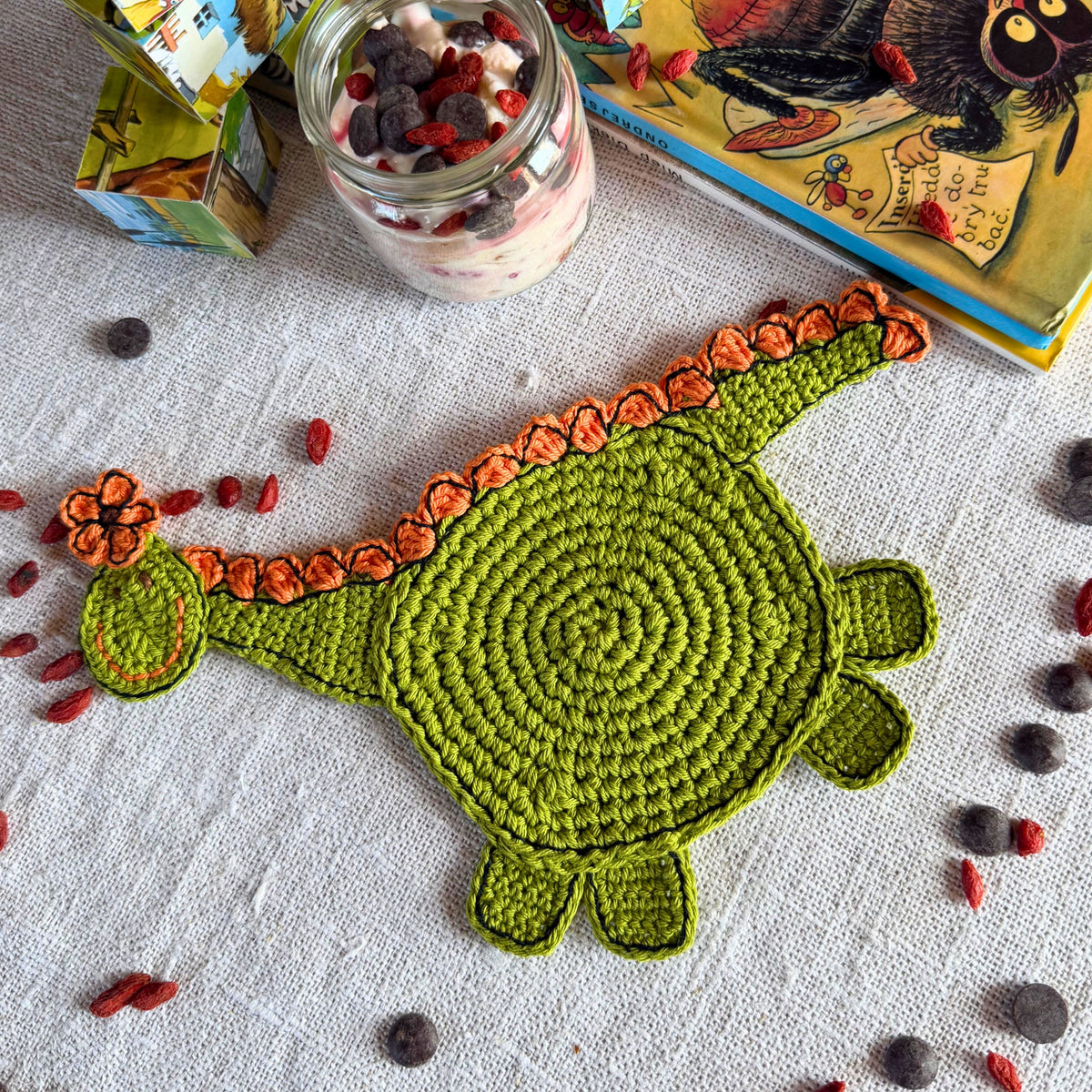 A crocheted dinosaur-shaped mat in green with orange scalloped edges, laid flat on a white textured cloth with scattered red and dark berries. A glass jar of mixed berries, a colorful book, and a small box are visible around it.