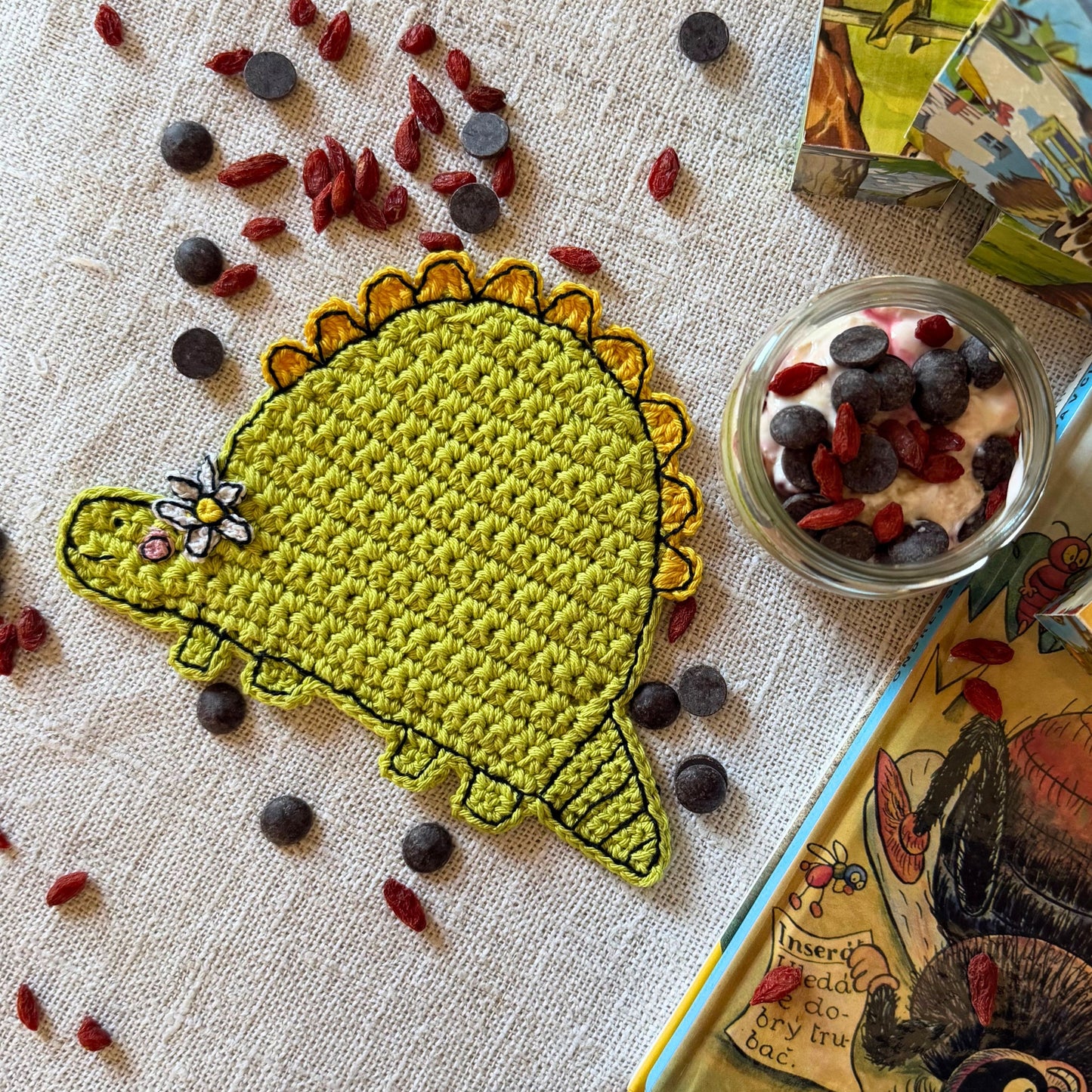 Green crochet dinosaur coaster with flower displayed beside a glass jar and illustrated book.