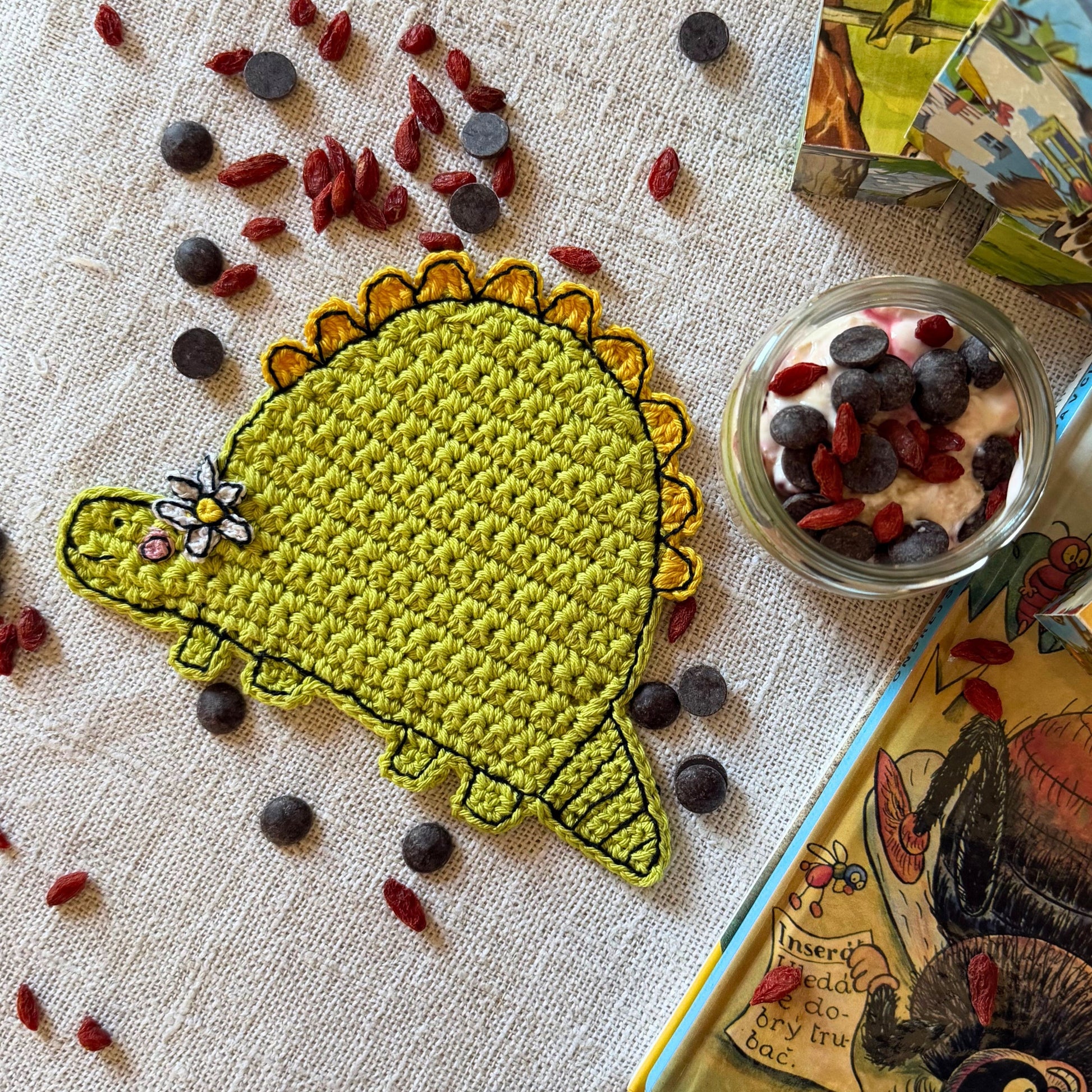 Green crochet dinosaur coaster with flower displayed beside a glass jar and illustrated book.