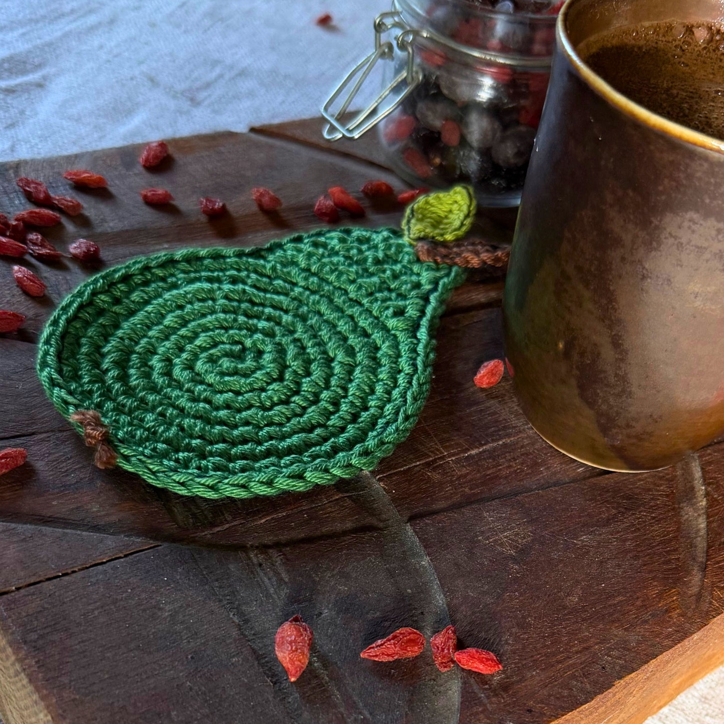 A green crocheted teardrop-shaped coaster or trivet with a small leaf detail, styled on a dark wooden surface with scattered red goji berries, a glass jar, and a copper mug.