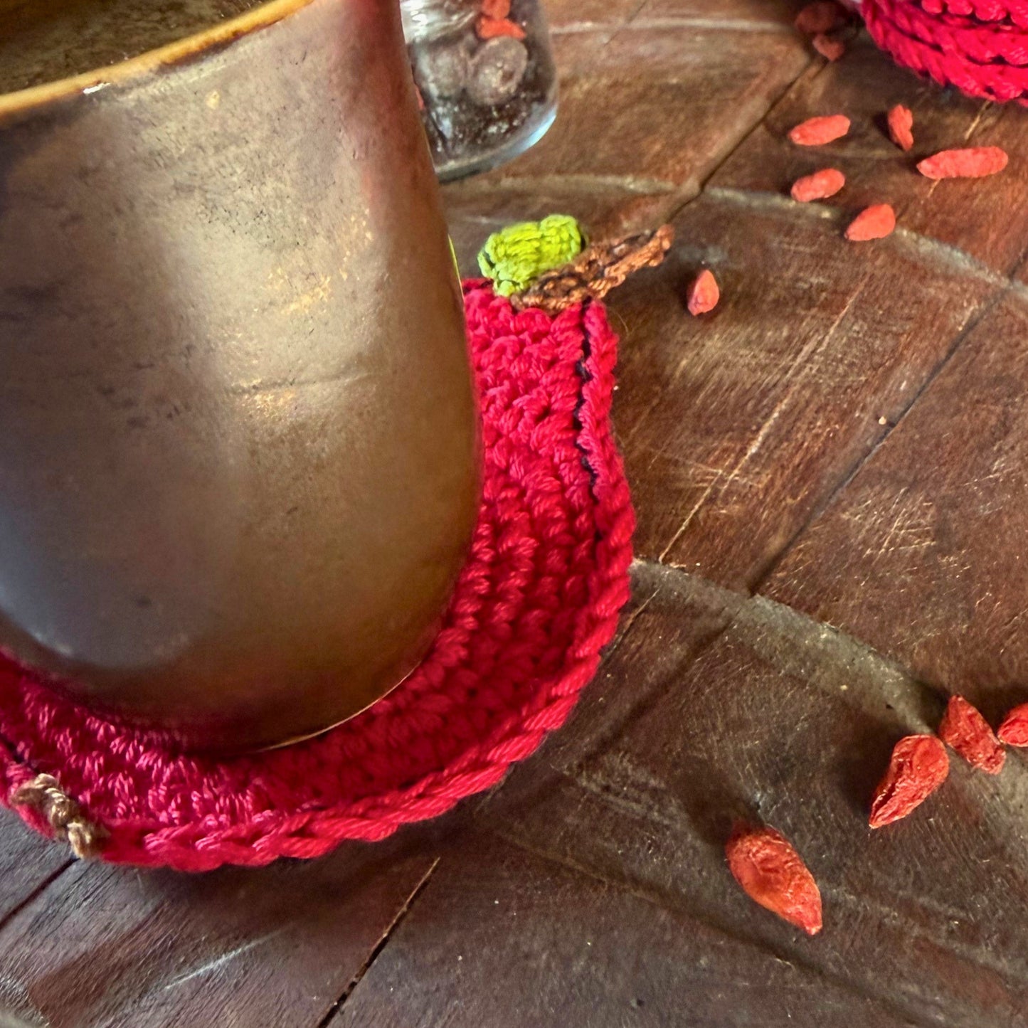 A close-up of a rustic mug resting on a bright red crocheted coaster with a small green and brown  detail, set on a dark wooden tabletop with scattered goji berries.