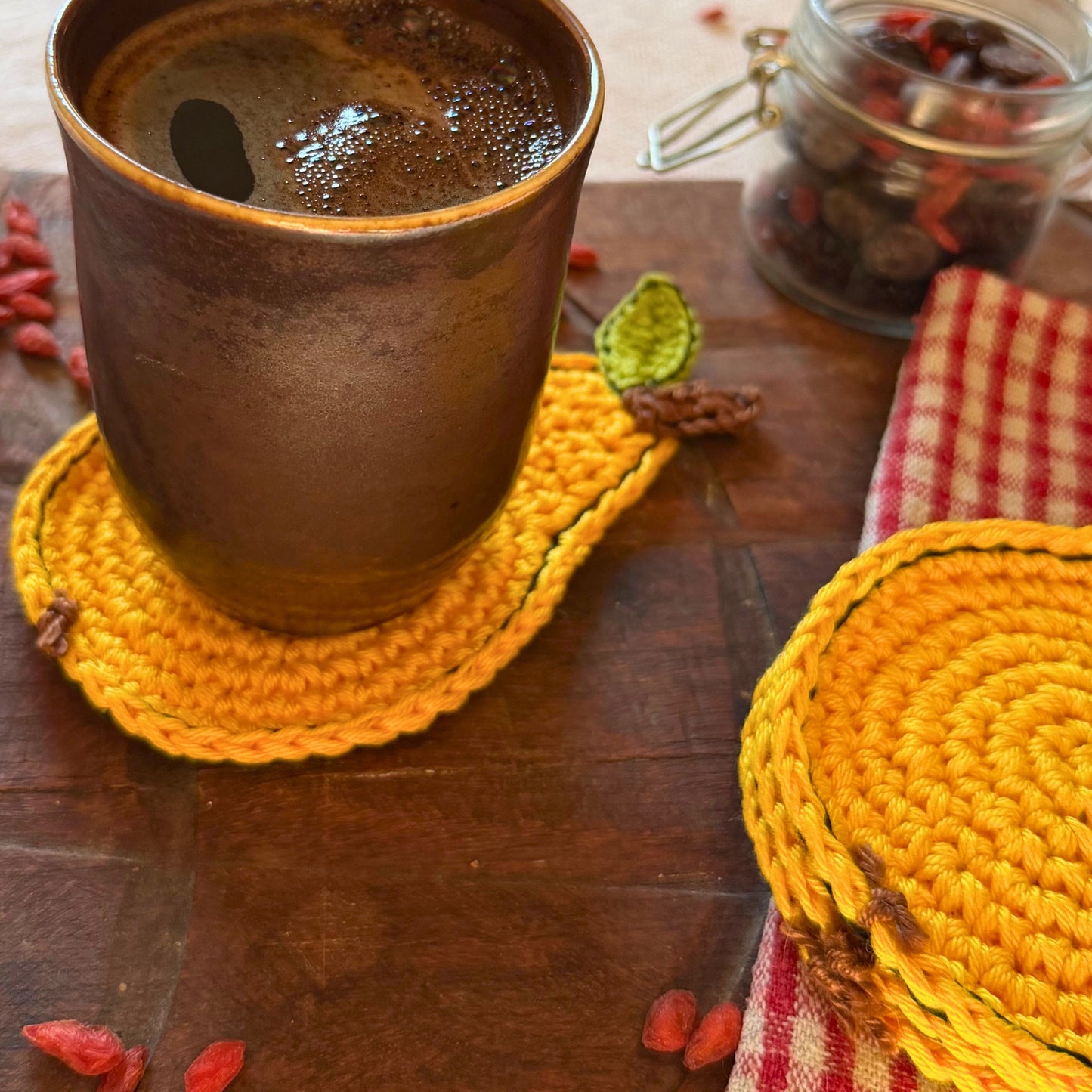 A rustic ceramic mug filled with a dark beverage sits on a yellow crocheted pear-shaped coaster on a wooden table. Another matching yellow coaster is visible on a red-and-white checkered cloth, with goji berries and a glass jar in the background.