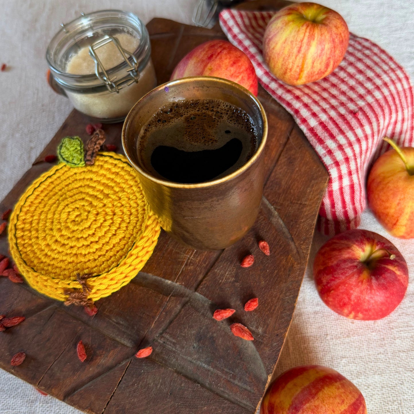 A styled flat-lay featuring a yellow hand-crocheted apple -shaped coasters on a rustic wooden board, next to a cup of black coffee and apples on a red checkered cloth.