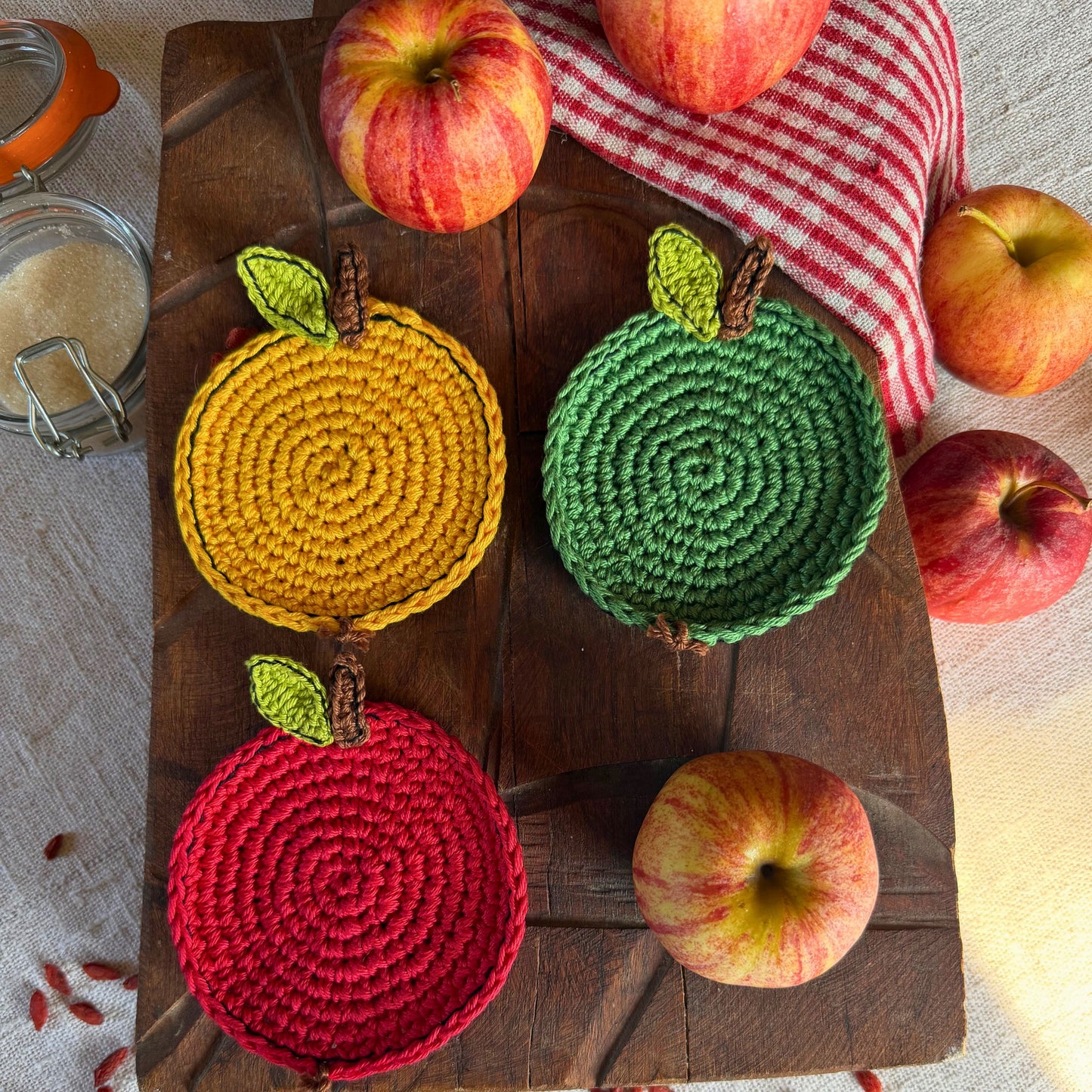 Three crocheted coasters shaped like apples in yellow, green, and red, arranged on a dark wooden board with whole apples and a red-and-white checkered cloth around them.