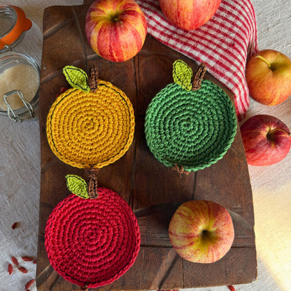 Three crocheted coasters shaped like apples in yellow, green, and red, arranged on a dark wooden board with whole apples and a red-and-white checkered cloth around them.