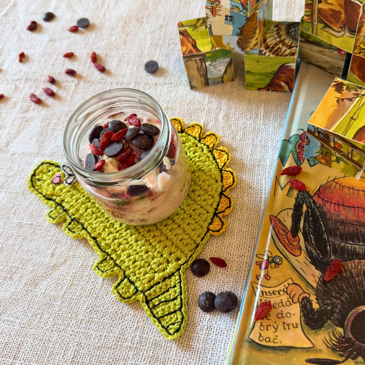 Green crochet dinosaur coaster used under a glass jar beside an illustrated book and decorative blocks.