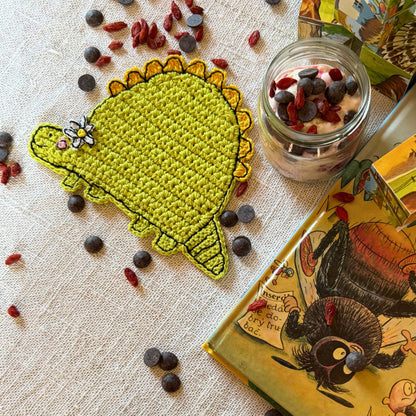 Green crochet dinosaur coaster with flower displayed on a linen table beside a glass jar and illustrated book.