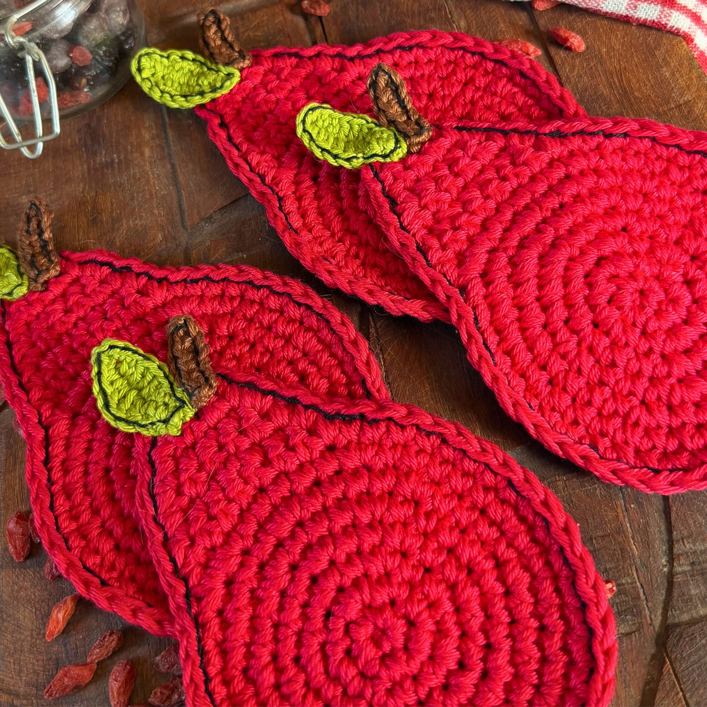 Four red crocheted apple-shaped coasters with green leaves and brown stems, arranged on a dark wooden tabletop.