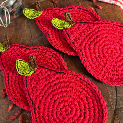 Four red crocheted apple-shaped coasters with green leaves and brown stems, arranged on a dark wooden tabletop.