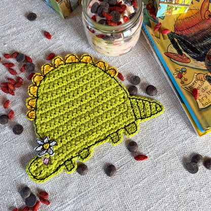 Green crochet dinosaur coaster with flower displayed on a linen table beside a glass jar and illustrated book.