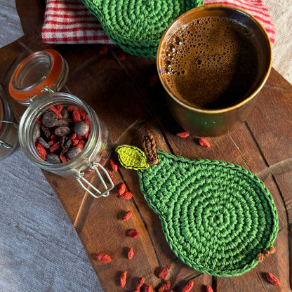 A green crocheted pear-shaped coaster or trivet with a leaf detail, styled on a rustic wooden table alongside a cup of coffee, a glass jar of dried fruit, and a red-and-white checkered cloth.