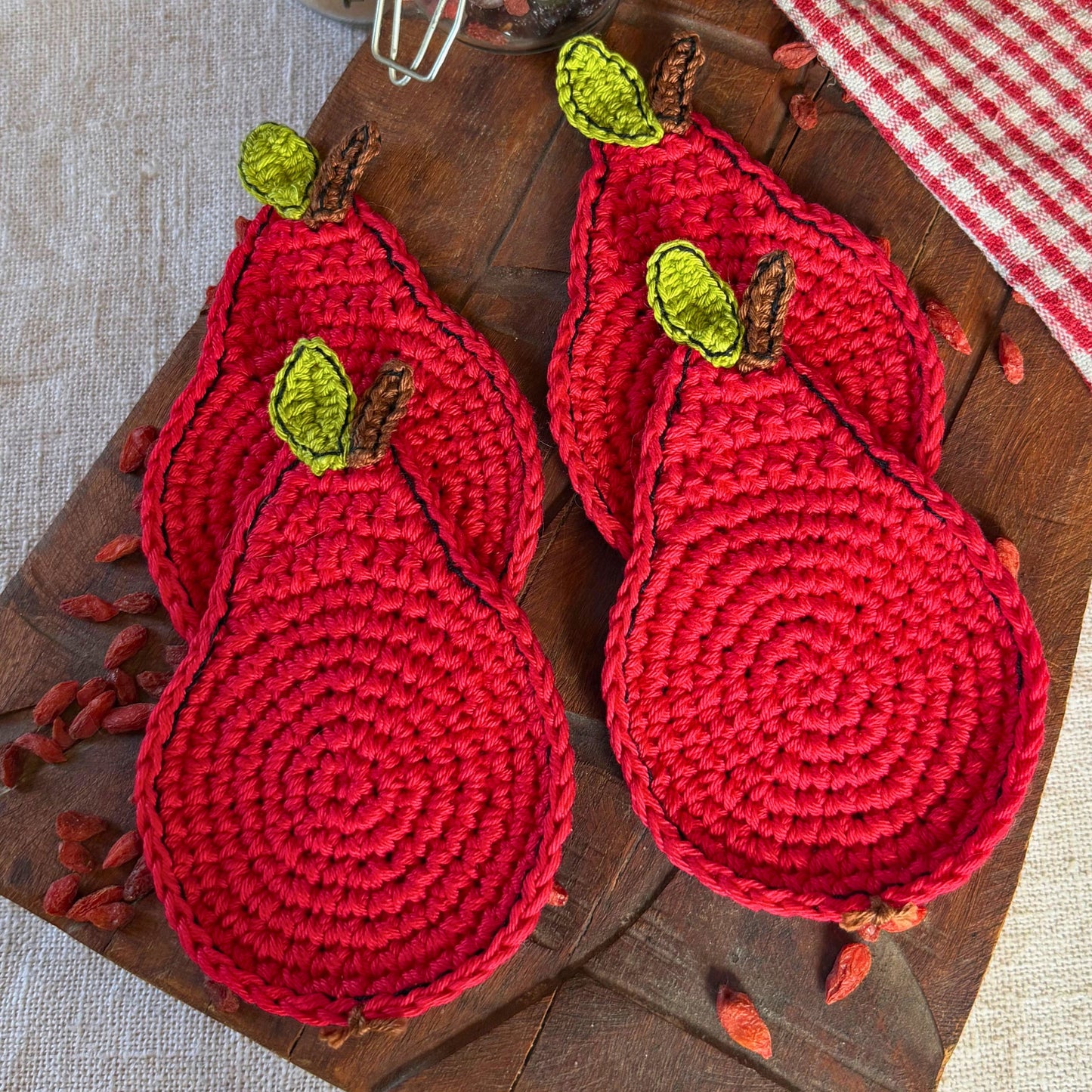 Four red crocheted pear-shaped coasters with green and brown leaf details, arranged on a rustic wooden board with scattered dried red berries.