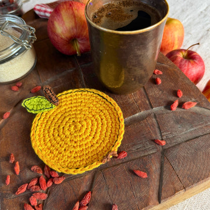 A round yellow crocheted coaster shaped like a apple, featuring a green leaf and brown stem, displayed on a rustic wooden board with apples, goji berries, and a cup of coffee.