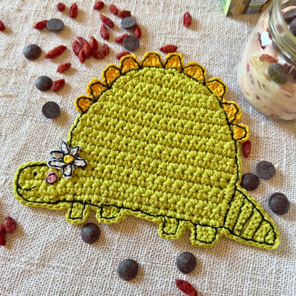 Close-up of a green crochet dinosaur coaster with yellow back details and a small white flower.