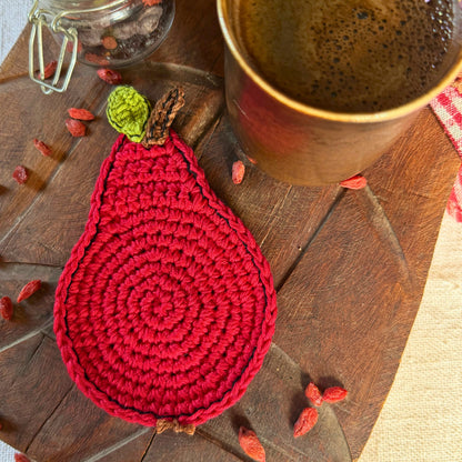 A red crocheted coaster shaped like a pear with a green top and brown stem, styled on a wooden surface next to a ceramic mug and dried red goji berries.