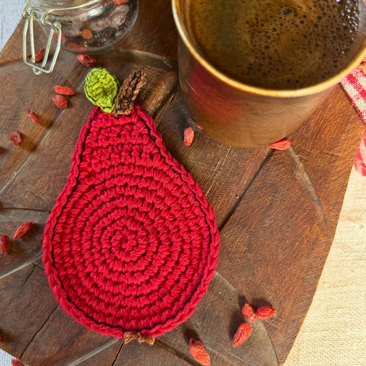 A red crocheted coaster shaped like a pear with a green top and brown stem, styled on a wooden surface next to a ceramic mug and dried red goji berries.