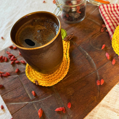 A dark brown ceramic mug filled with coffee sits on a yellow crocheted coaster on a rustic wooden board, surrounded by scattered red goji berries and a cozy red-and-white checkered cloth.