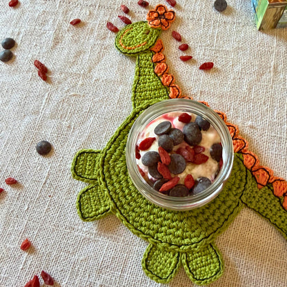 A green crocheted dinosaur-shaped coaster with orange accents, holding a small glass jar filled with yogurt and topped with blueberries and red goji berries, set on a beige linen surface with scattered red berries.