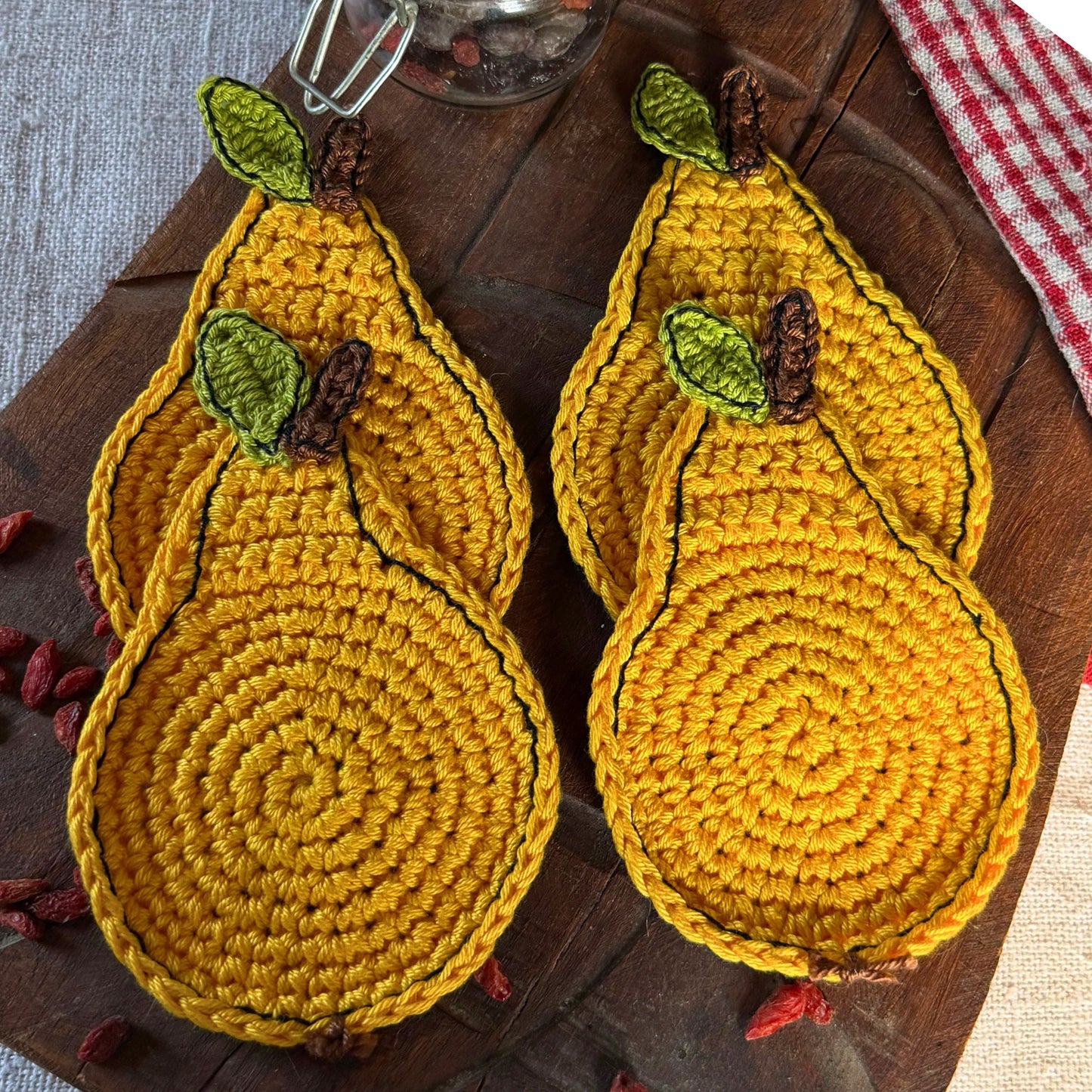 Four yellow crocheted pear-shaped coasters with green leaves and brown stems, arranged on a dark wooden serving board with a red-and-white checkered cloth.