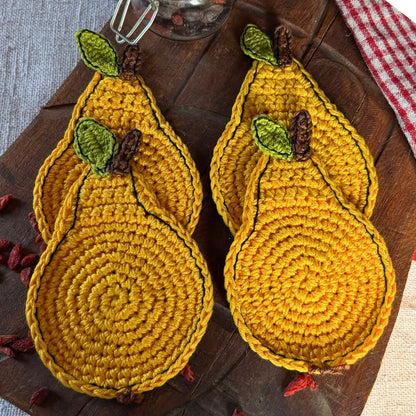 Four yellow crocheted pear-shaped coasters with green leaves and brown stems, arranged on a dark wooden serving board with a red-and-white checkered cloth.