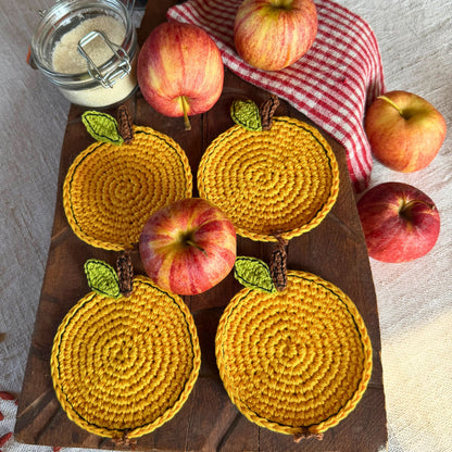 Four round yellow crocheted coasters with green leaf accents and brown stem details, arranged on a dark wooden board with whole apples and a red-and-white checkered cloth.