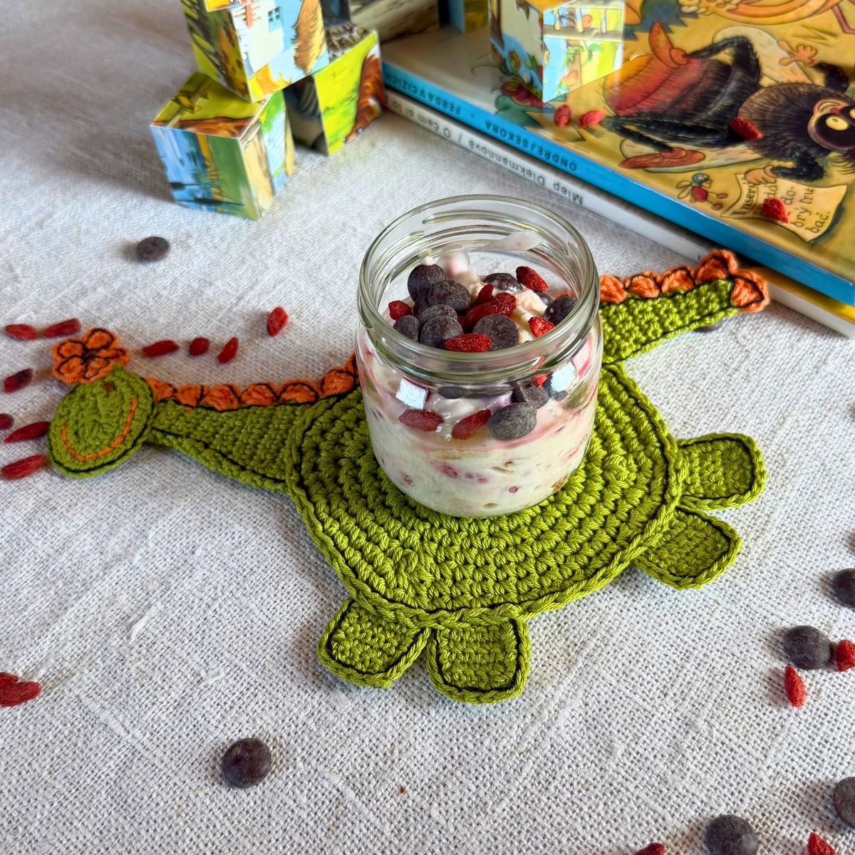 A small glass jar filled with layered yogurt, berries, and fruit sits on a green crocheted dinosaur-shaped coaster on a white textured tablecloth. Books and colorful blocks are blurred in the background.