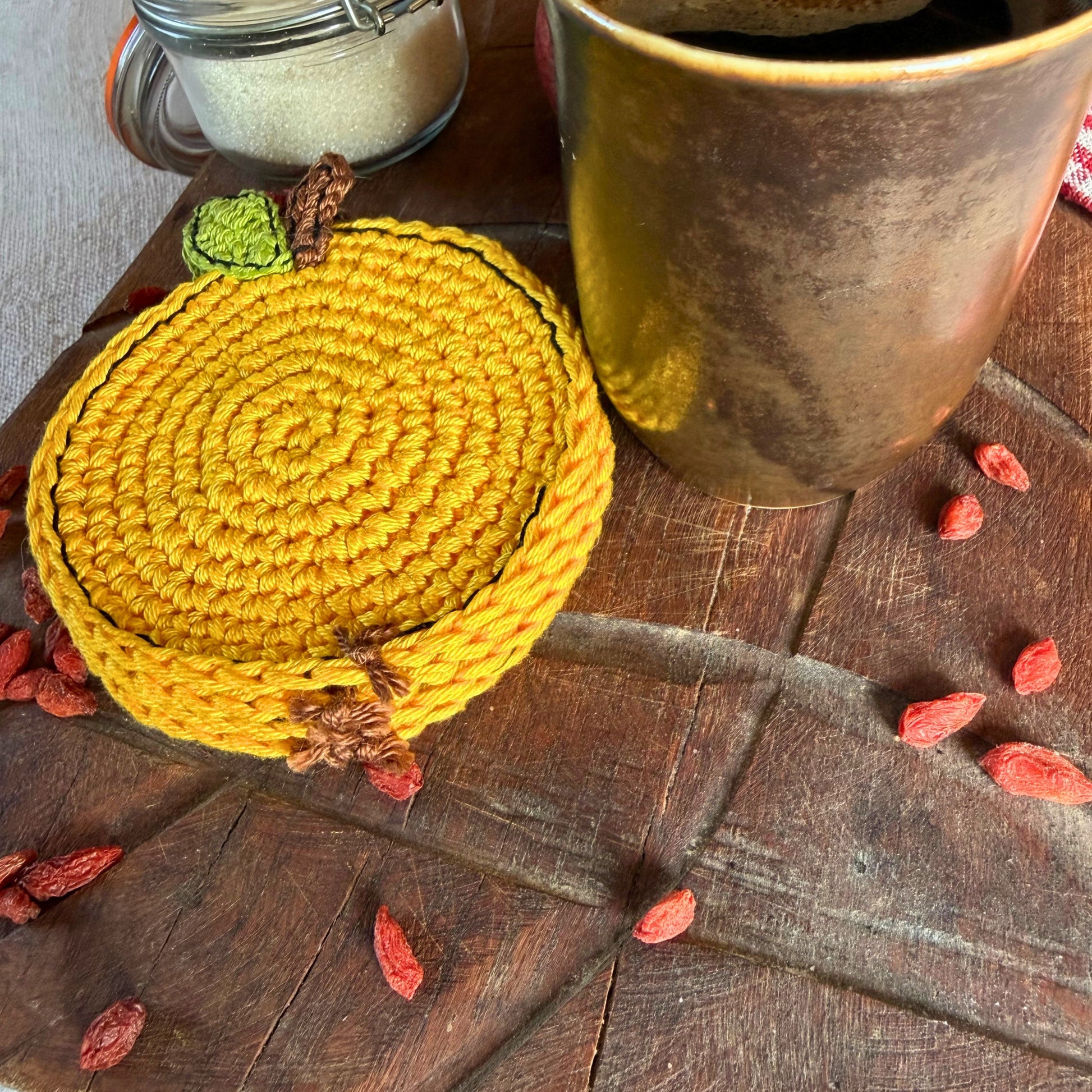 A round yellow crocheted coasters with a small green and brown stem detail, styled on a rustic dark wooden table next to a ceramic mug and scattered red goji berries.