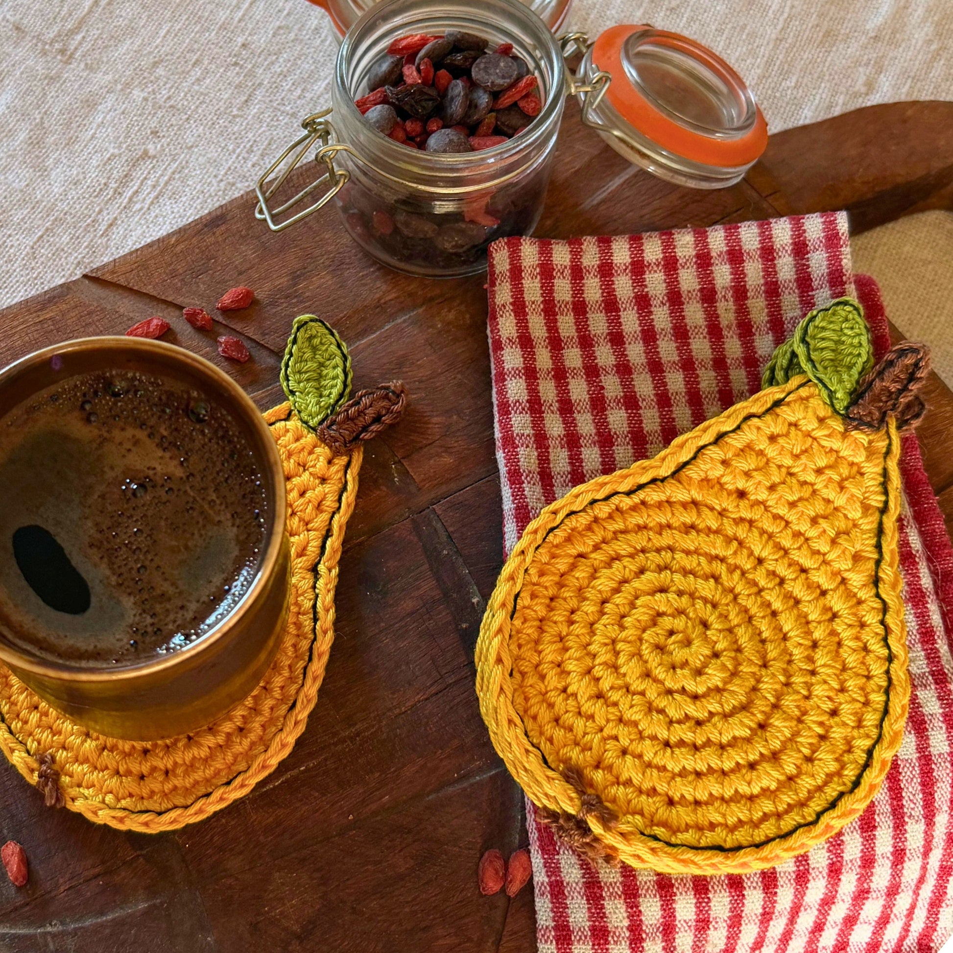 A set of crocheted pear-shaped coasters  in warm yellow , styled on a rustic wooden table with a cup of coffee, a red-and-white checkered napkin, and a glass jar of dried fruit.