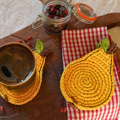 A set of crocheted pear-shaped coasters  in warm yellow , styled on a rustic wooden table with a cup of coffee, a red-and-white checkered napkin, and a glass jar of dried fruit.