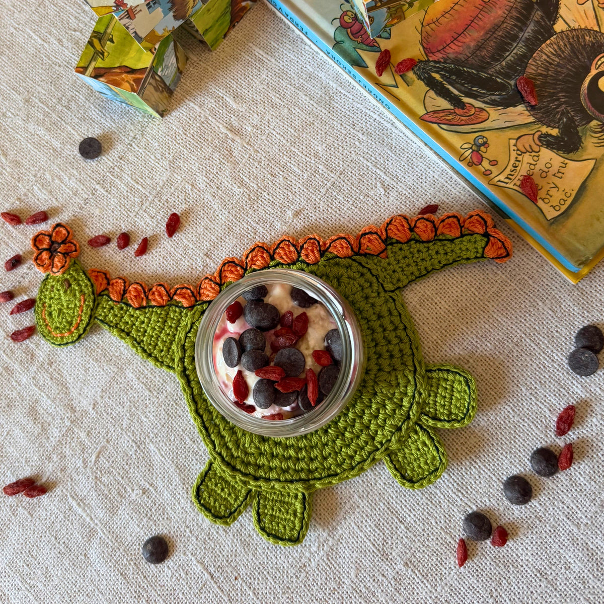 A crocheted dinosaur-shaped jar coaster in green and orange, holding a small glass jar filled with yogurt and berries, styled on a textured cream linen surface with scattered dried berries and children's books in the background.
