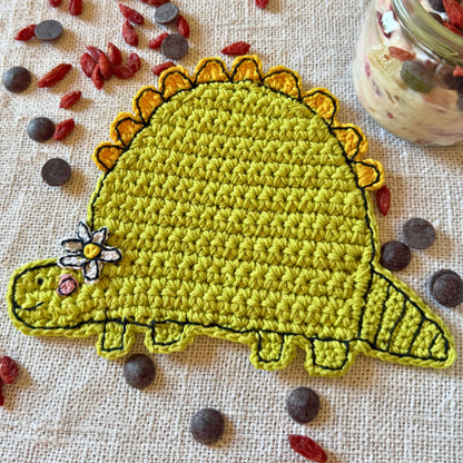 Green crochet dinosaur coaster with flower displayed on a linen table beside a glass jar.
