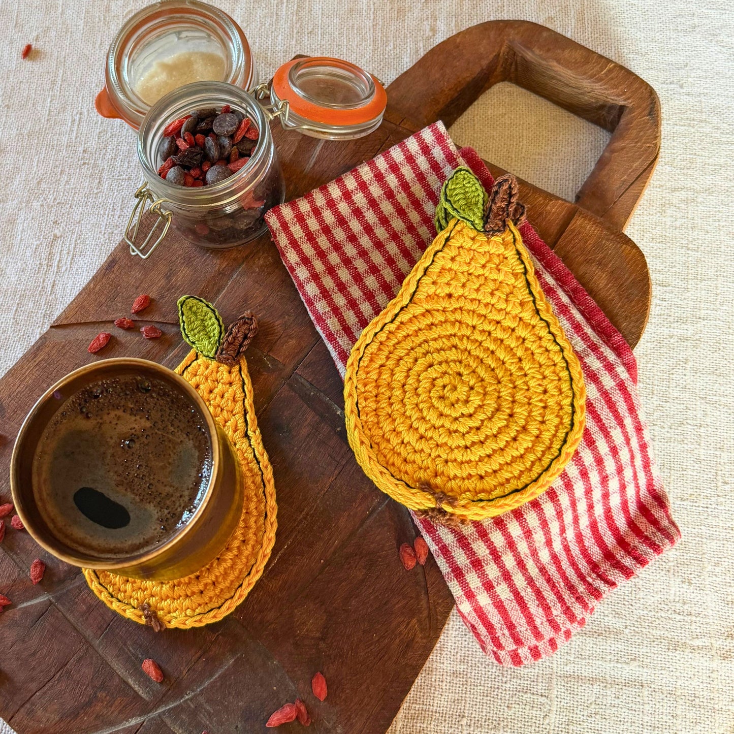 Two crocheted pear-shaped coasters in mustard yellow and red-and-white gingham fabric, styled on a wooden board with a cup of coffee and small glass jars on a linen surface.