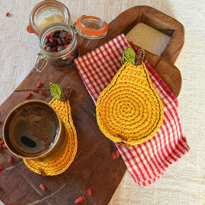 Two crocheted pear-shaped coasters in mustard yellow and red-and-white gingham fabric, styled on a wooden board with a cup of coffee and small glass jars on a linen surface.