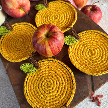 Four yellow crocheted apple coasters with green leaves and brown stems, arranged on a wooden board with whole apples as styling props.