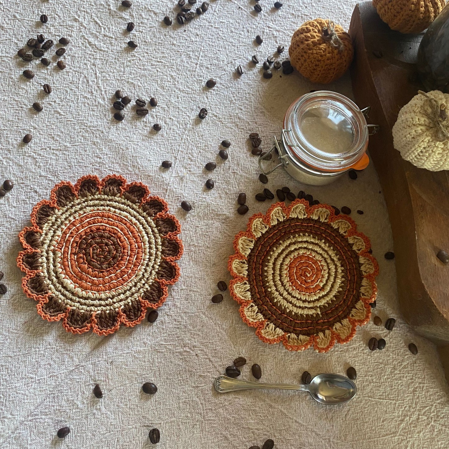 Two crocheted coasters with a warm color scheme on a textured surface with coffee beans and a jar.