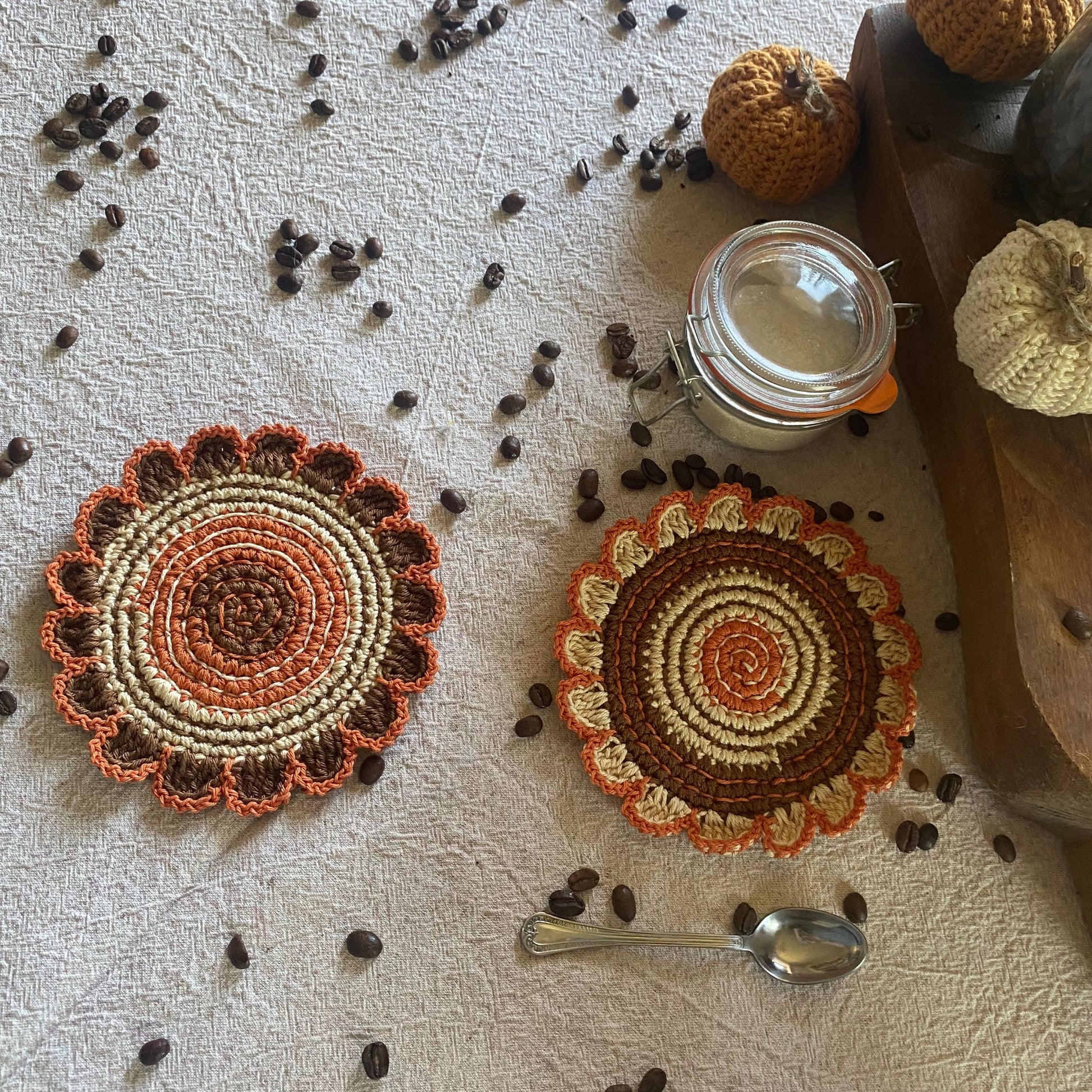 Two crocheted coasters with a warm color scheme on a textured surface with coffee beans and a jar.