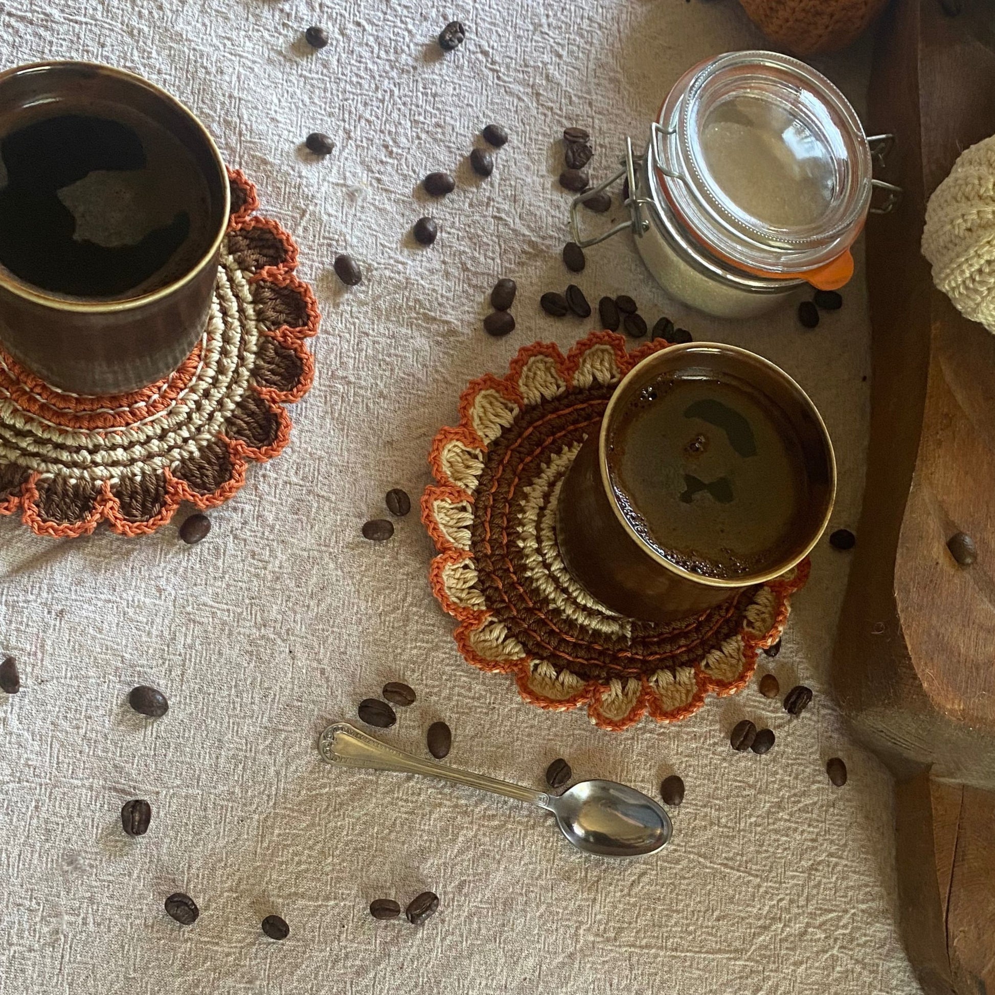 Two cups of coffee on crocheted coasters with scattered coffee beans on a textured surface.