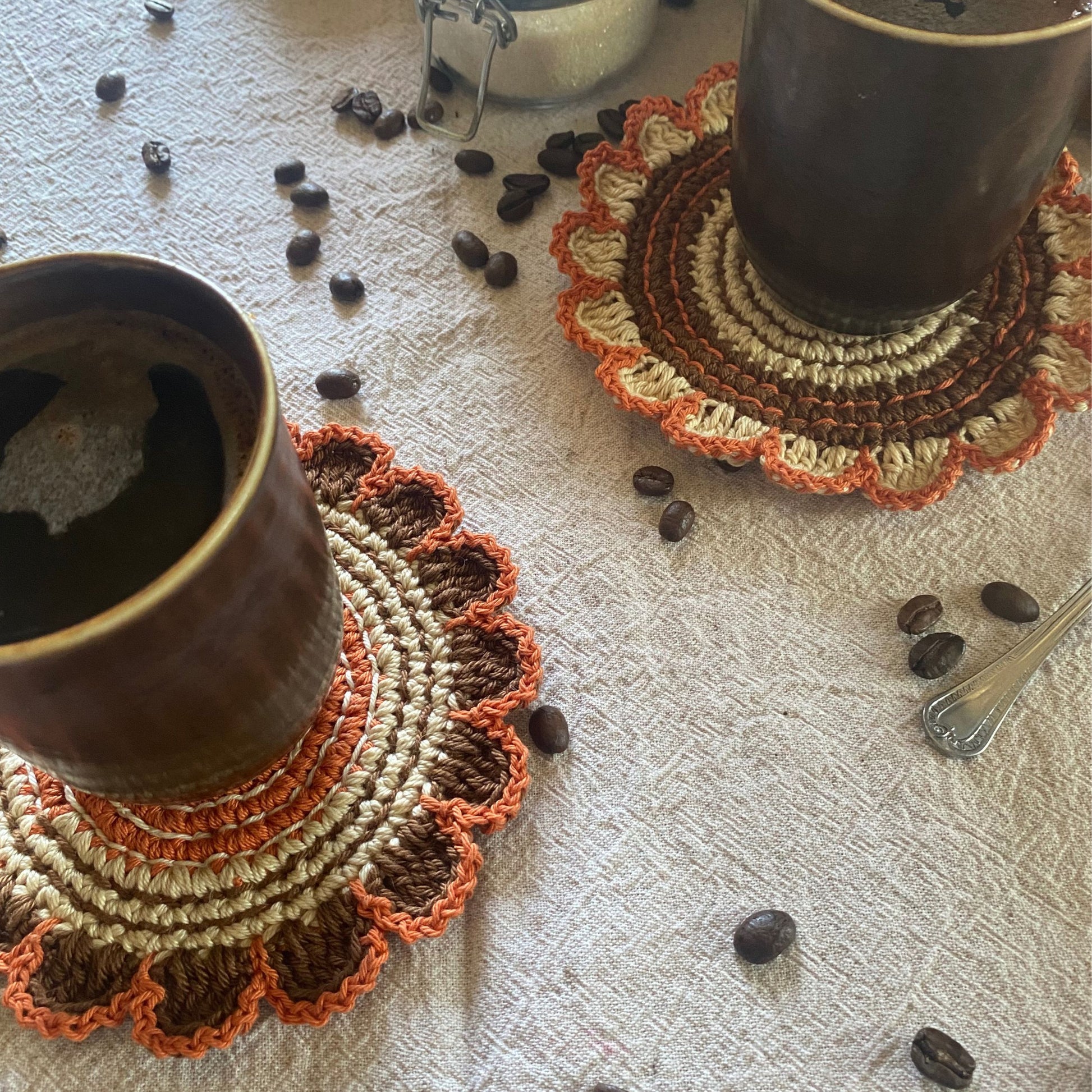 Two ceramic cups on crocheted coasters with scattered coffee beans on a textured surface.