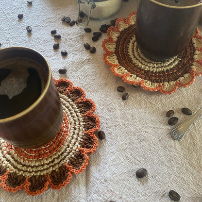 Two ceramic cups on crocheted coasters with scattered coffee beans on a textured surface.