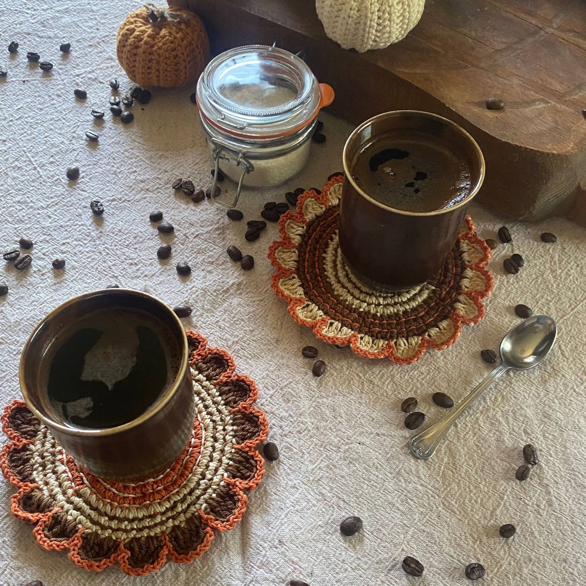Two cups of coffee on crocheted coasters with scattered coffee beans and a jar in the background.