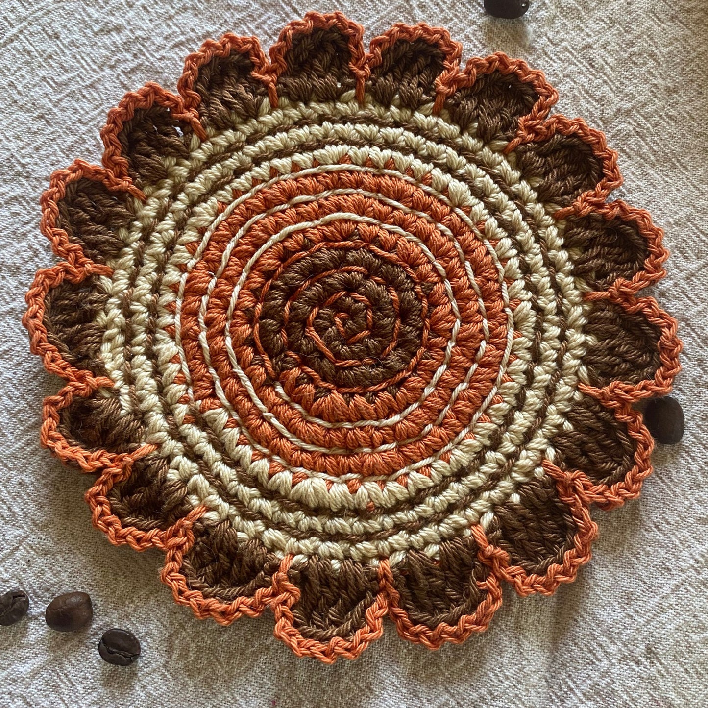 Round crochet mat with concentric circle pattern in brown, orange, and beige on a textured surface.
